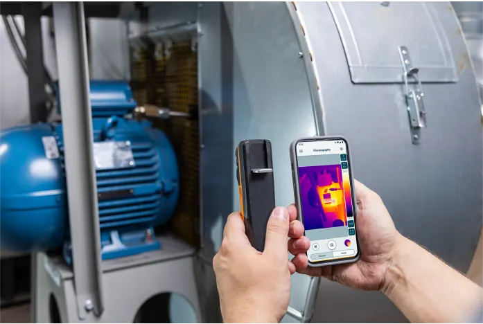 Person using a smartphone with thermal imaging app to inspect machinery.