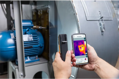Person using a smartphone with thermal imaging app to inspect machinery.